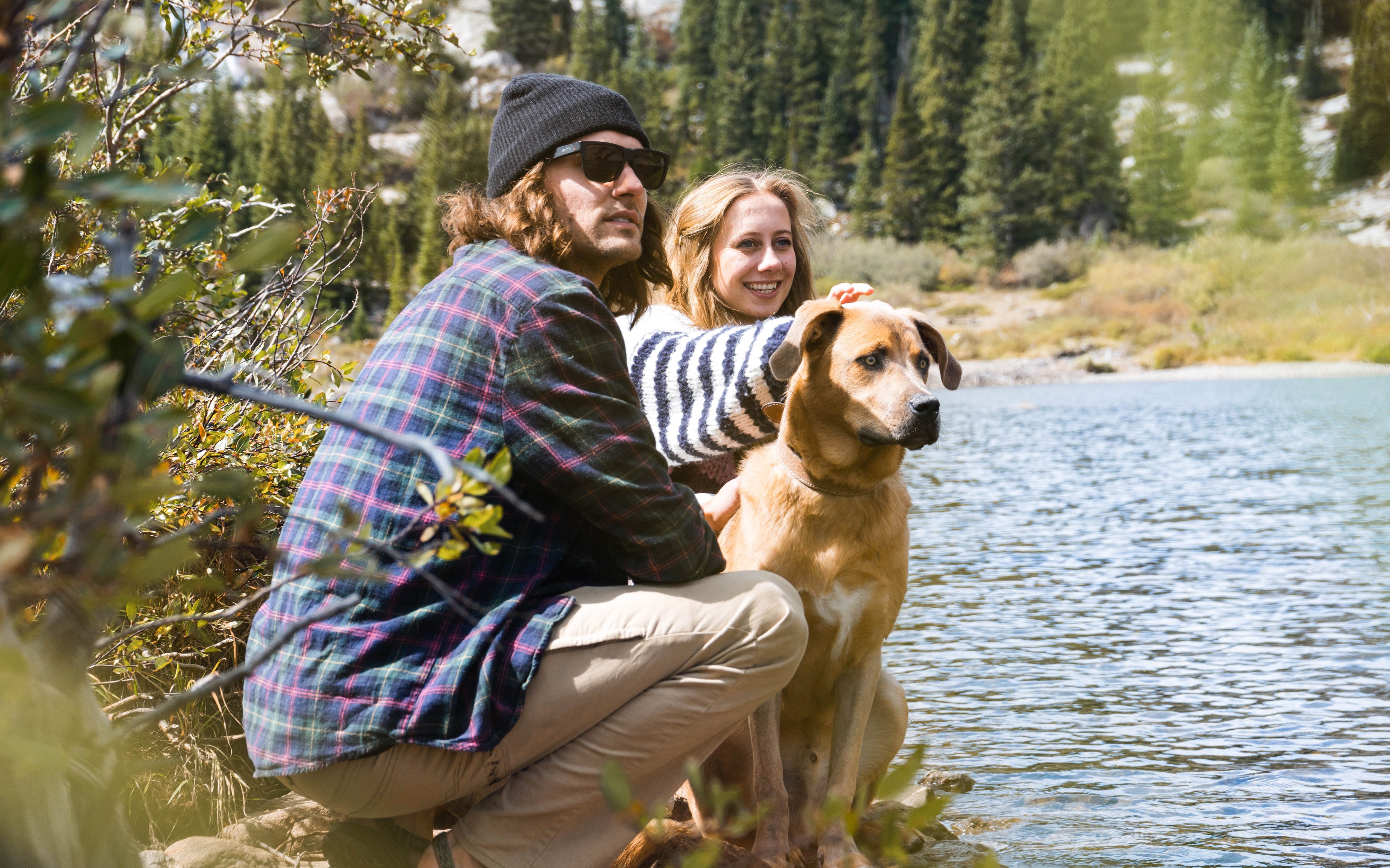 Man with a beanie and woman in striped sweater with a large tan dog sitting by the water in nature